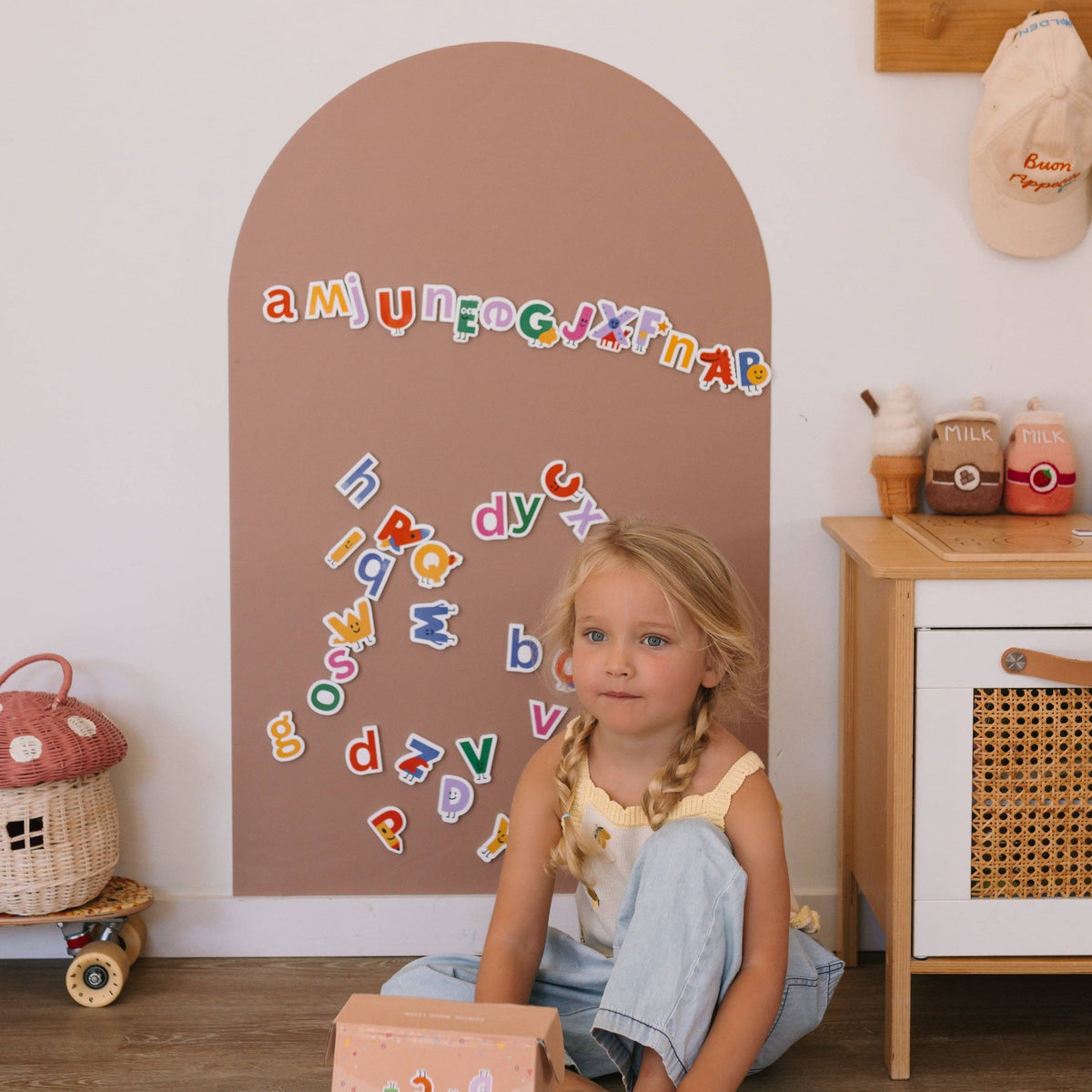 A young girl with blonde pigtails sits on the floor in a child's playroom, looking at the camera. In front of her is a large arched brown wall plaque decorated with colorful alphabet letters.