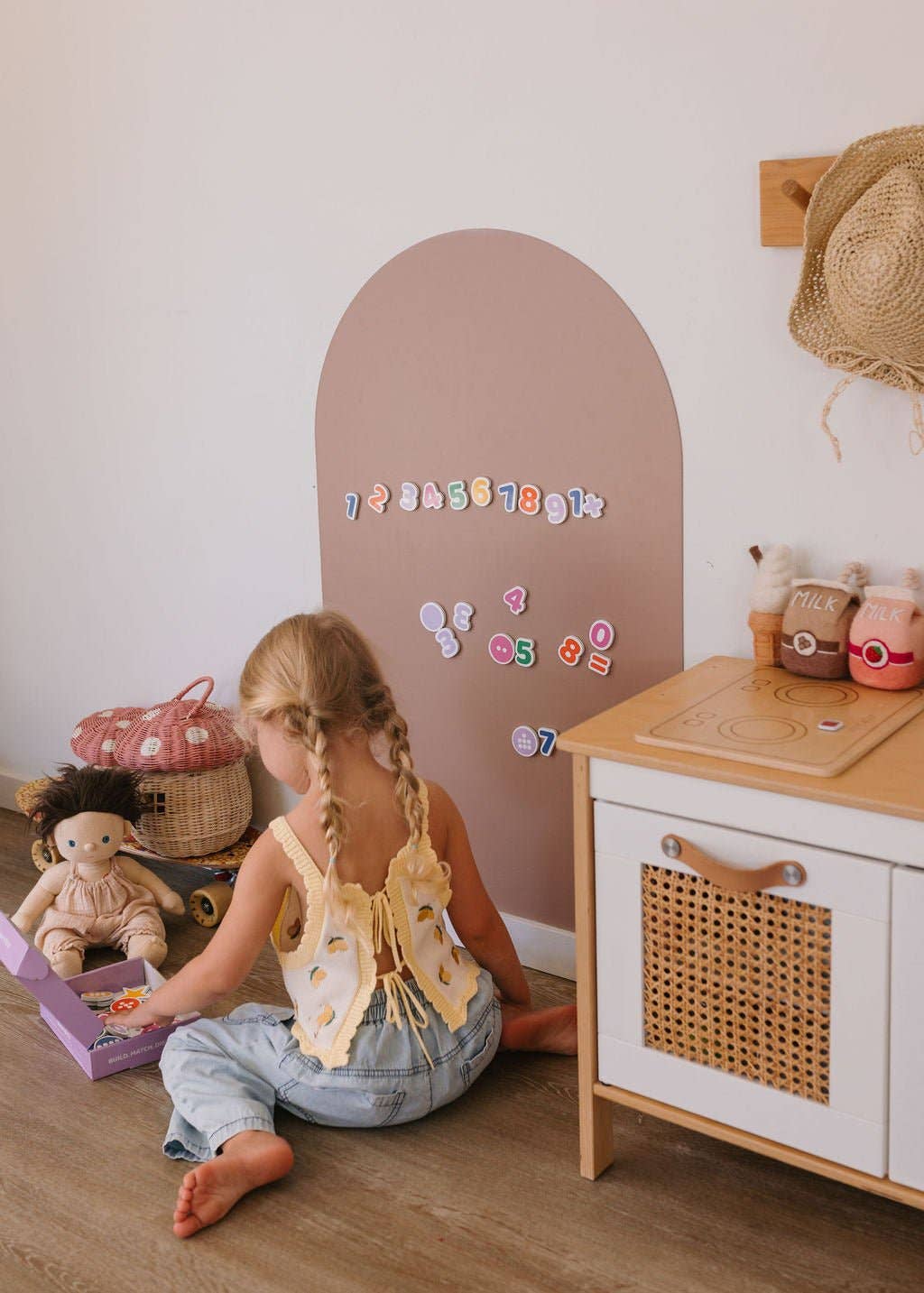A young girl with braided hair sits on the floor in a child's playroom, wearing a yellow butterfly-wing backless top and light blue denim jeans. She is interacting with toys near a wooden play kitchen, a pink arch-shaped wall decoration with colorful magnetic numbers, and a straw hat hanging on the wall.