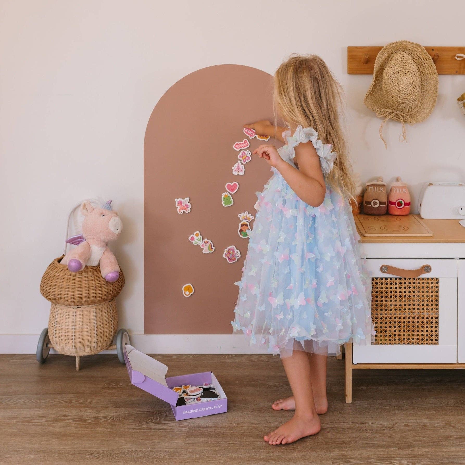 Young girl in a polka dot dress playing with stickers in a room with toys and a mirror.