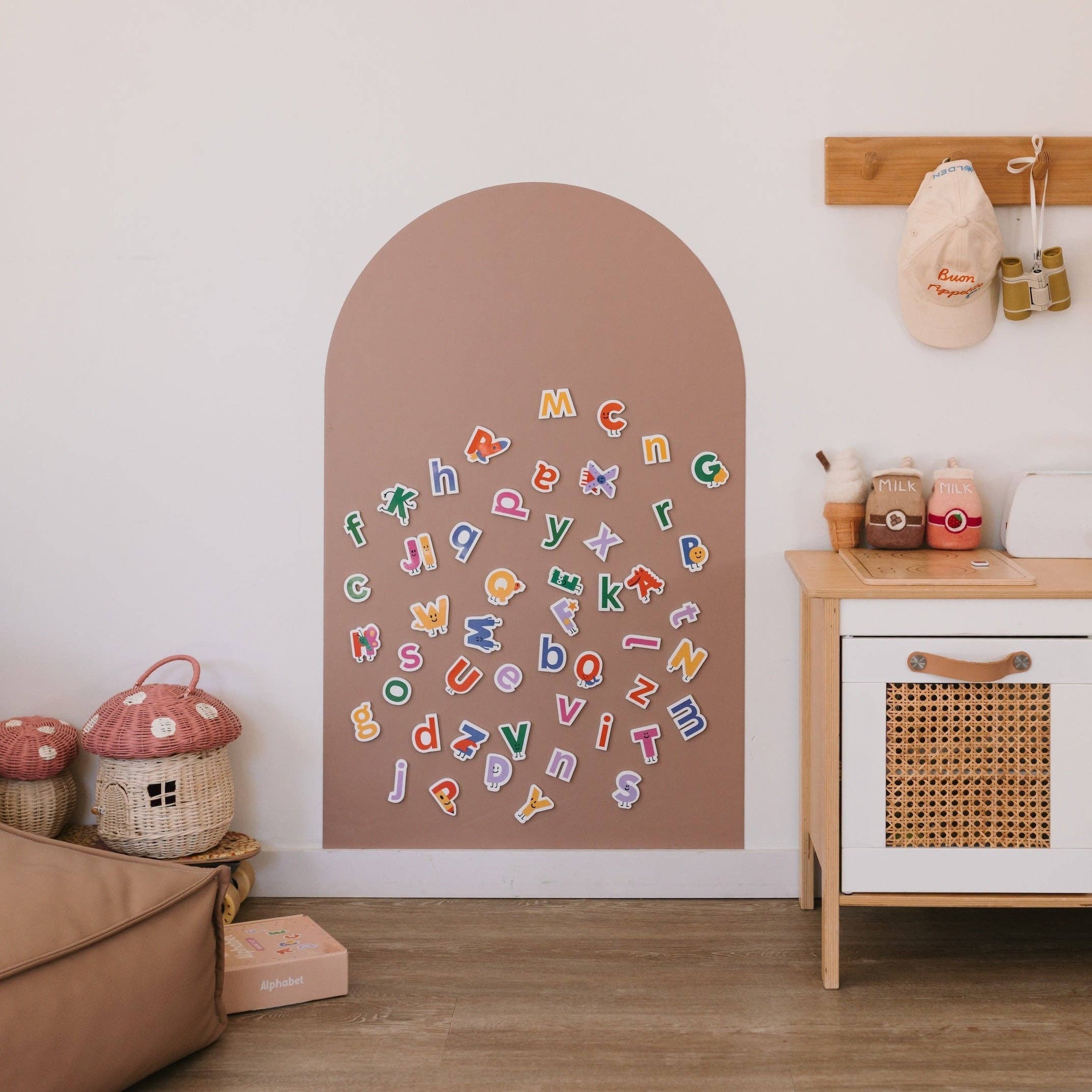 A styled children's room corner featuring a large arch-shaped brown magnetic wall board on the wall, covered with colorful alphabet letters. To the right is a light wood and white cabinet with a woven front, and to the left are mushroom-shaped baskets and a brown sofa.