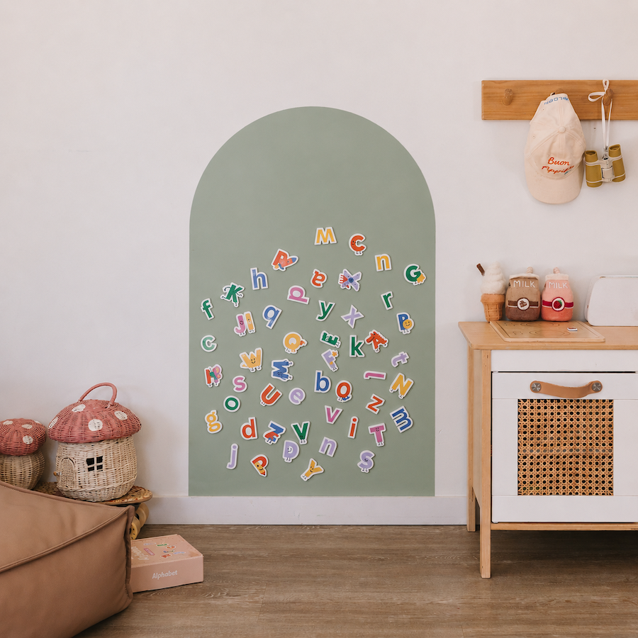 A styled children's room corner showing a sage green arched wall panel with colorful alphabet letters scattered on it. The room includes a wooden toy cabinet, mushroom-shaped baskets, a hat rack with a white cap, and a light wood floor.