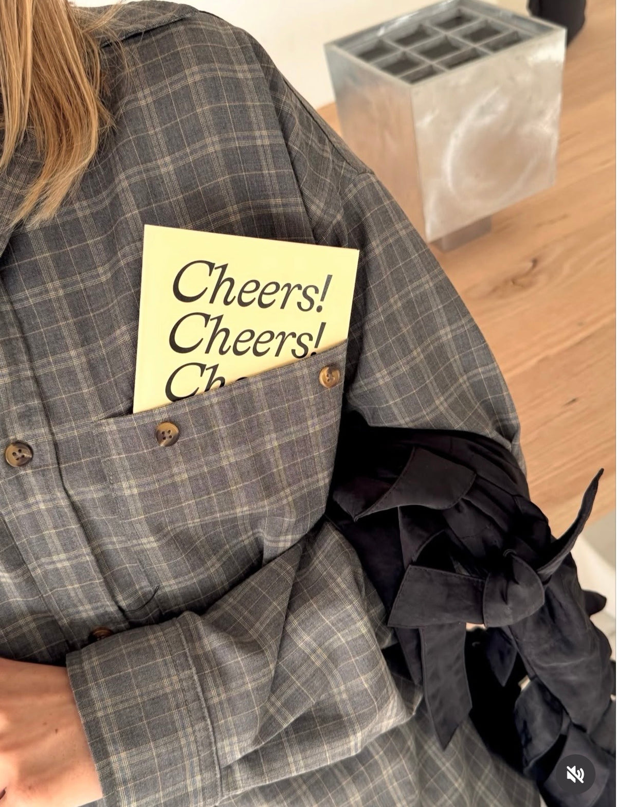 A close-up lifestyle shot of a person wearing a gray and black plaid button-up shirt with a pocket. A yellow sticky note reading 'Cheers! Cheers! Cheers!' is tucked into the pocket, and a black ruffled accessory is draped over the arm.