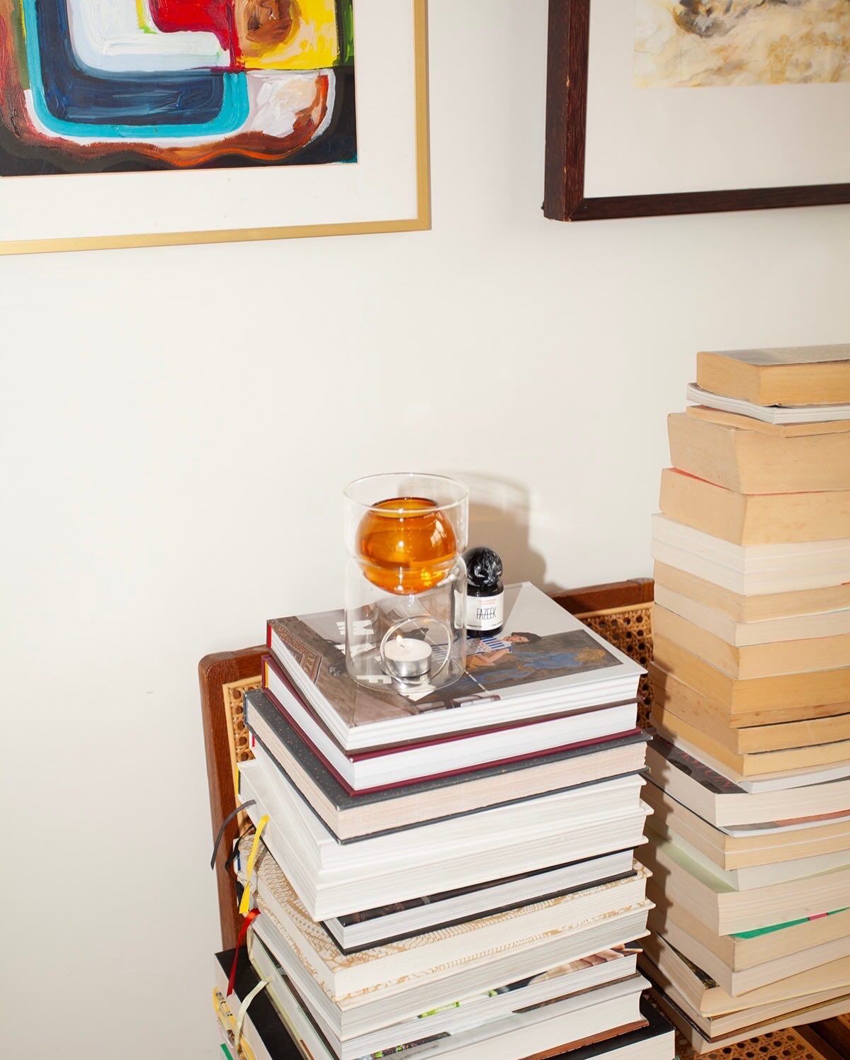 A clear glass tumbler with an amber liquid sits on top of a tall stack of books, alongside a small black bottle, in a styled interior setting with more books and framed wall art.