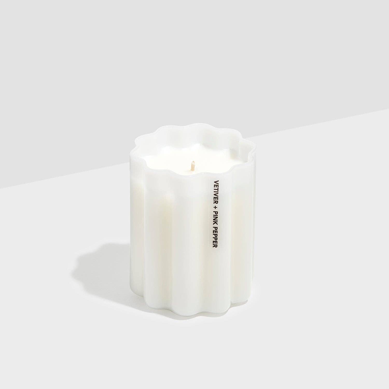 A white fluted candle with a scalloped top edge and a single wick, featuring vertical black text that reads 'VETTERIS + PINK PINK' on the side, set against a minimalist light gray background.