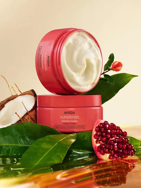 A jar of Aveda Nutrienish Hair Moisturizer shown open with white cream inside, stacked on top of another jar, surrounded by green leaves, a halved coconut, and a pomegranate on a warm beige background.