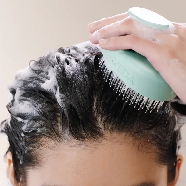 Person using a green comb to massage shampoo into their hair with a beige background