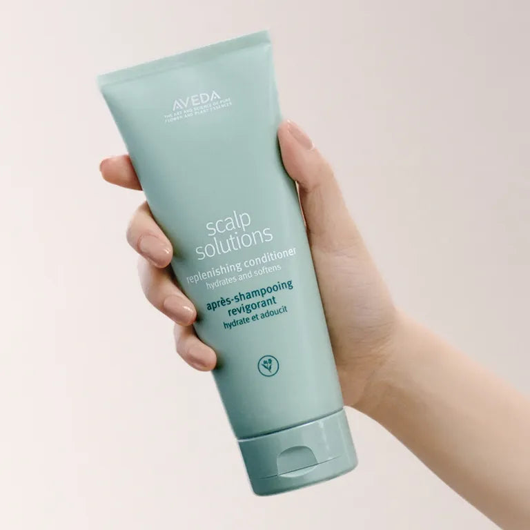 A hand holding a light blue-green squeeze tube of Aveda Scalp Solutions Replenishing Conditioner against a soft, pale pink background.