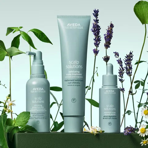 Three Aveda Scalp Solutions hair care products in a coordinated pale blue-gray set, arranged on a dark green pedestal with botanical props including lavender, mint, and white flowers against a soft mint background.
