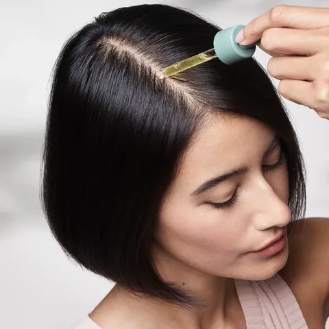 A close-up of a woman with a side part, showing a dropper applying a golden hair oil to her scalp. She has very dark, glossy black hair and is wearing a light pink top against a soft, light gray background.
