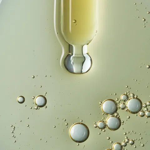 A close-up of a clear dropper or pipette tip containing pale yellow liquid, with a large transparent droplet hanging above a smooth pale greenish-beige surface dotted with small bubbles.