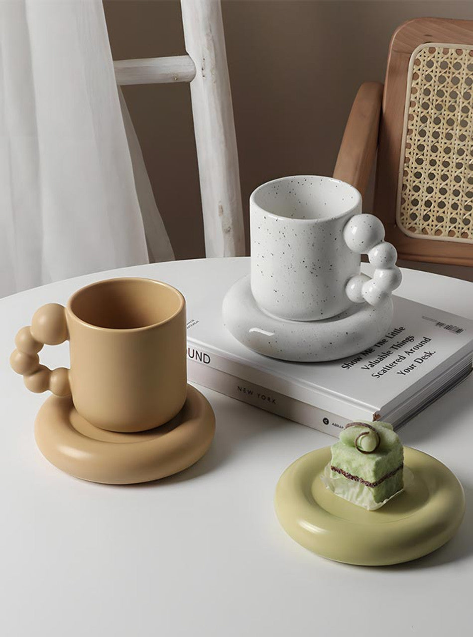A styled tabletop scene with ceramic mugs and matching saucers. In the foreground, a tan speckled mug sits on a matching round saucer with a bubble handle.