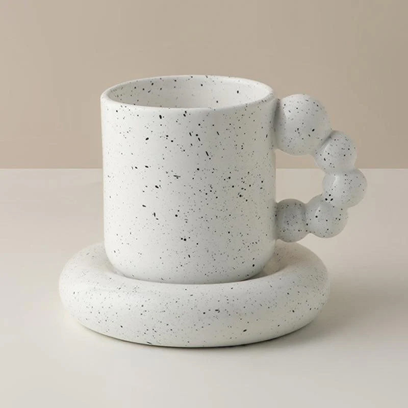A white speckled ceramic mug with a unique knotted handle, resting on a matching round saucer against a neutral beige background.