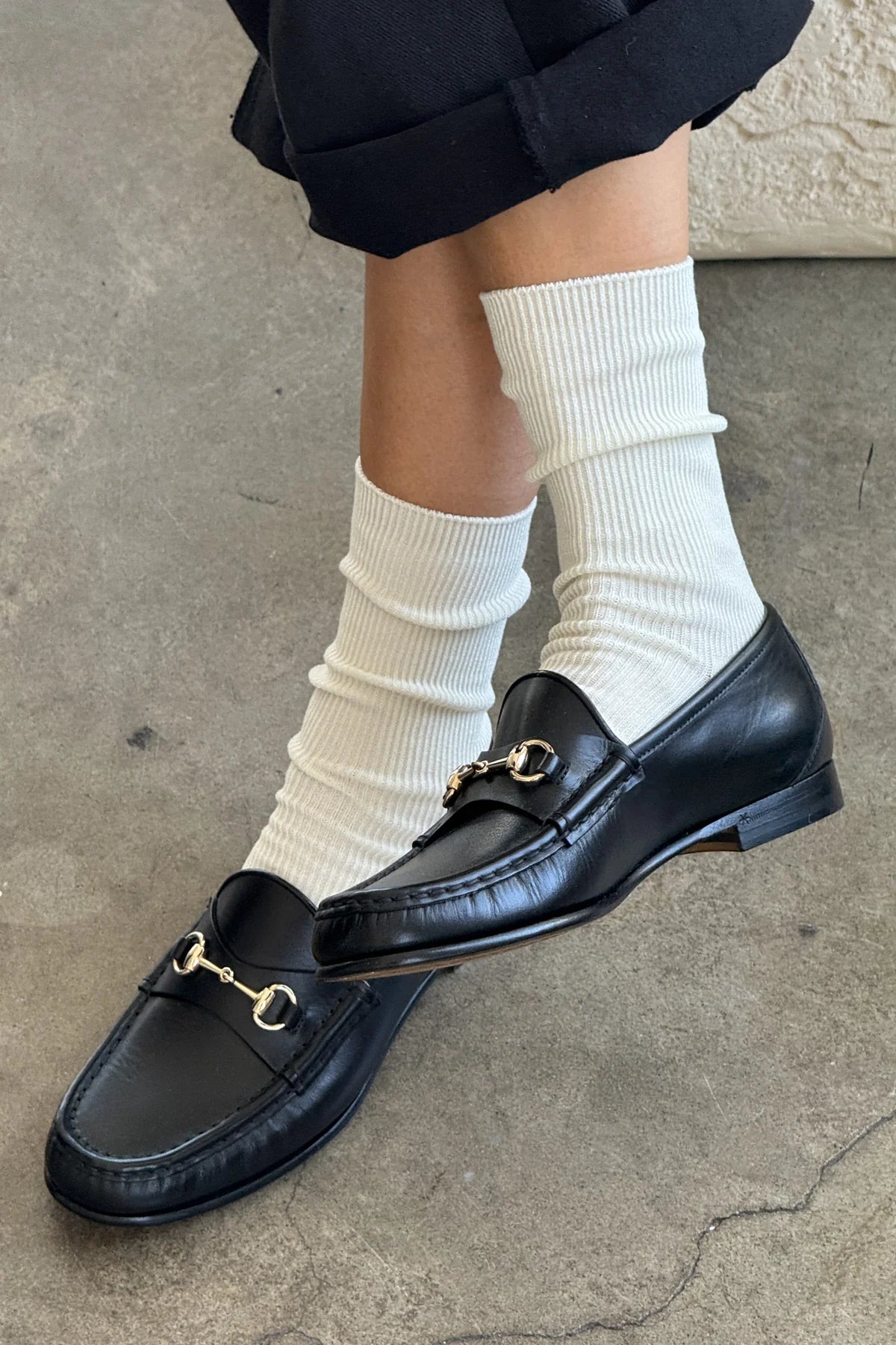 Black loafers with gold buckles worn with white socks on a concrete floor.