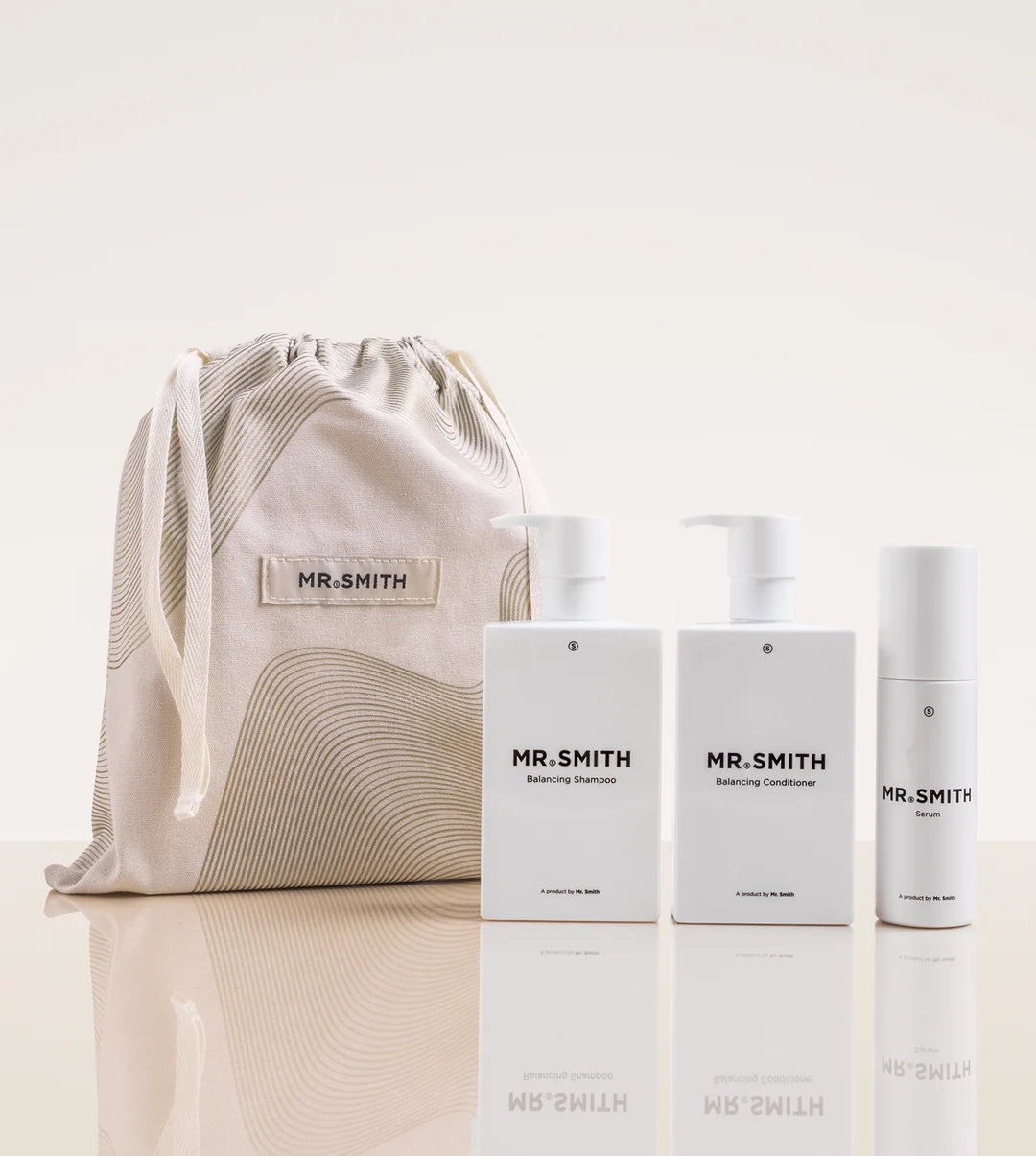 A Mr. Smith skincare gift set featuring three white bottles (Exfoliating Cleanser, Purifying Mask, and Deep Moisturizer) alongside a beige drawstring pouch with a matching label, displayed on a reflective cream surface.