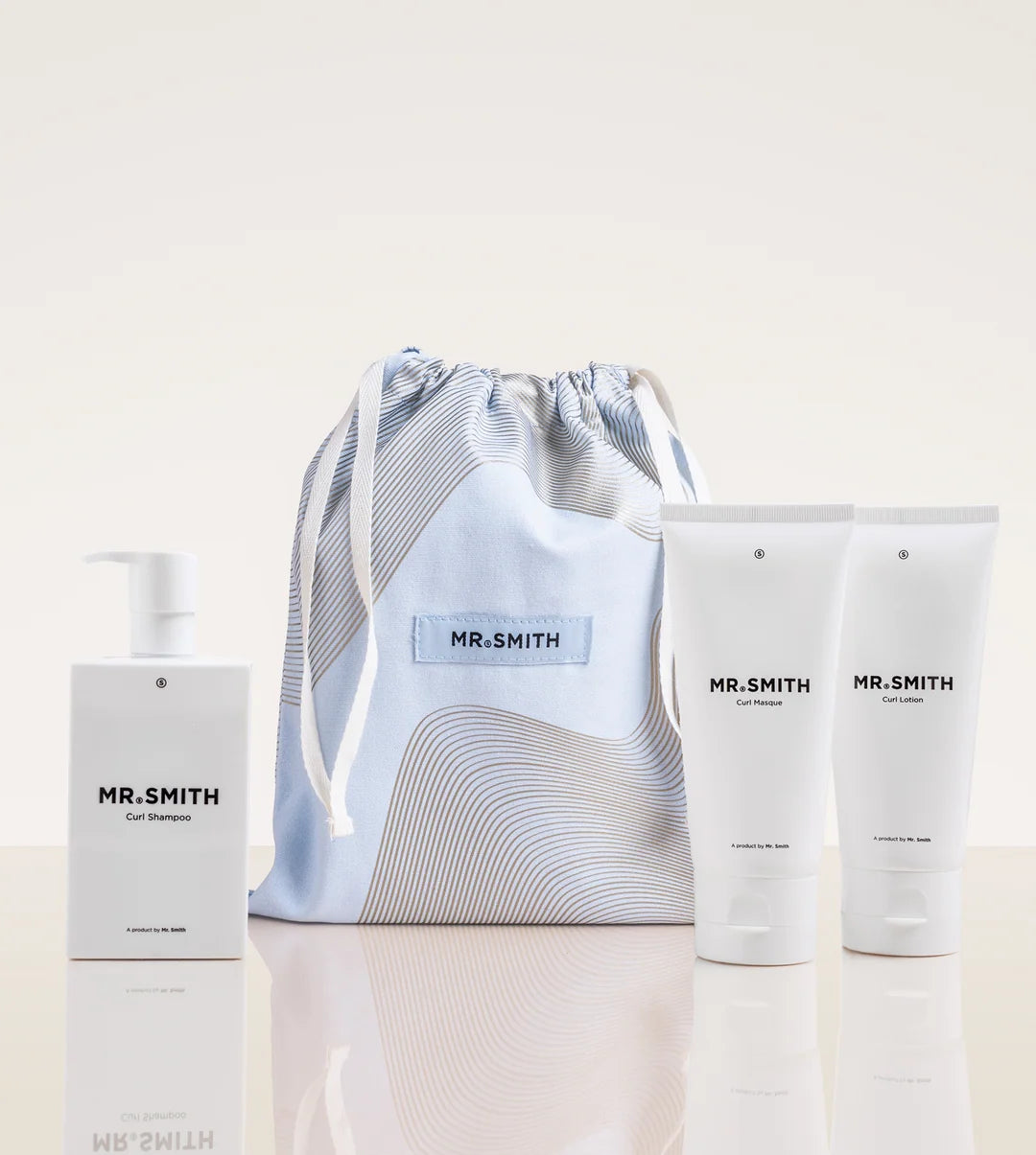 A Mr. Smith toiletry gift set featuring a white Full Shampoo bottle, a light blue and white striped drawstring pouch with a blue label, and two white body wash tubes, all on a reflective white surface against a warm off-white background.