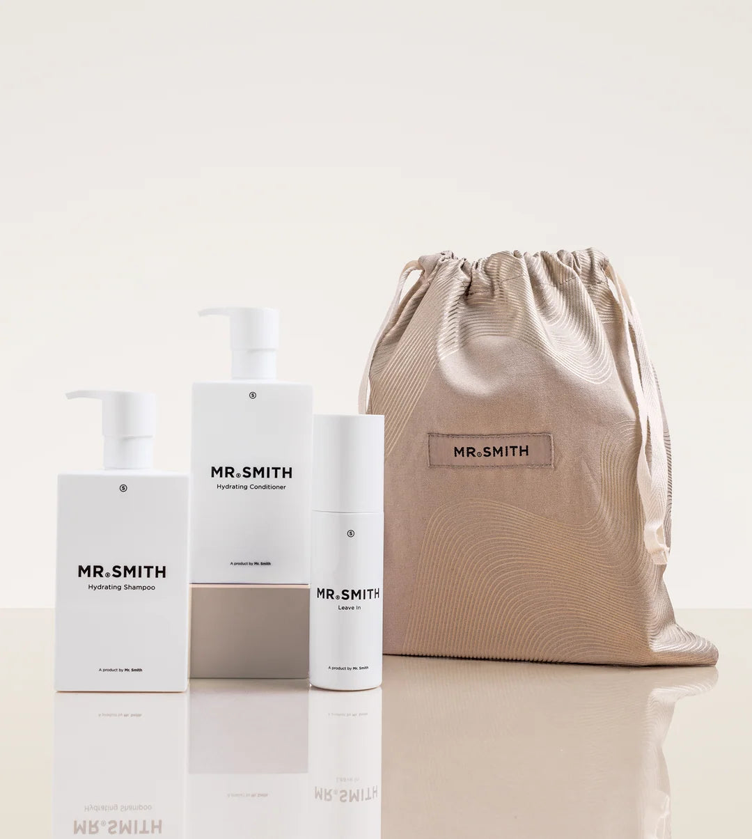 A Mr. Smith haircare gift set featuring a white shampoo bottle, a white conditioner box, a white cylindrical bottle, and a metallic beige drawstring pouch with a Mr.