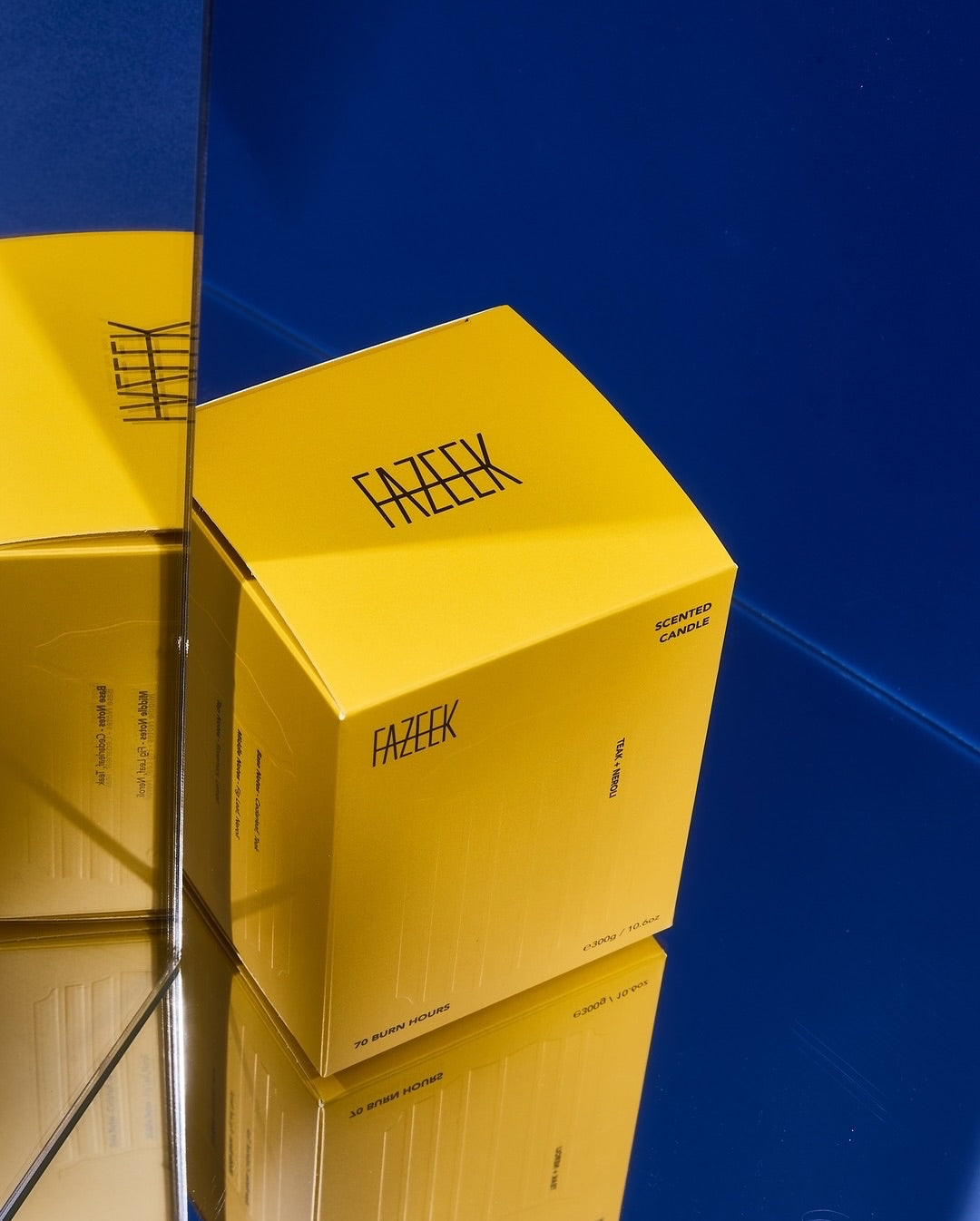 A bright yellow rectangular product box with the brand name 'FAZEEK' printed in black on multiple sides, photographed on a glossy blue reflective surface.