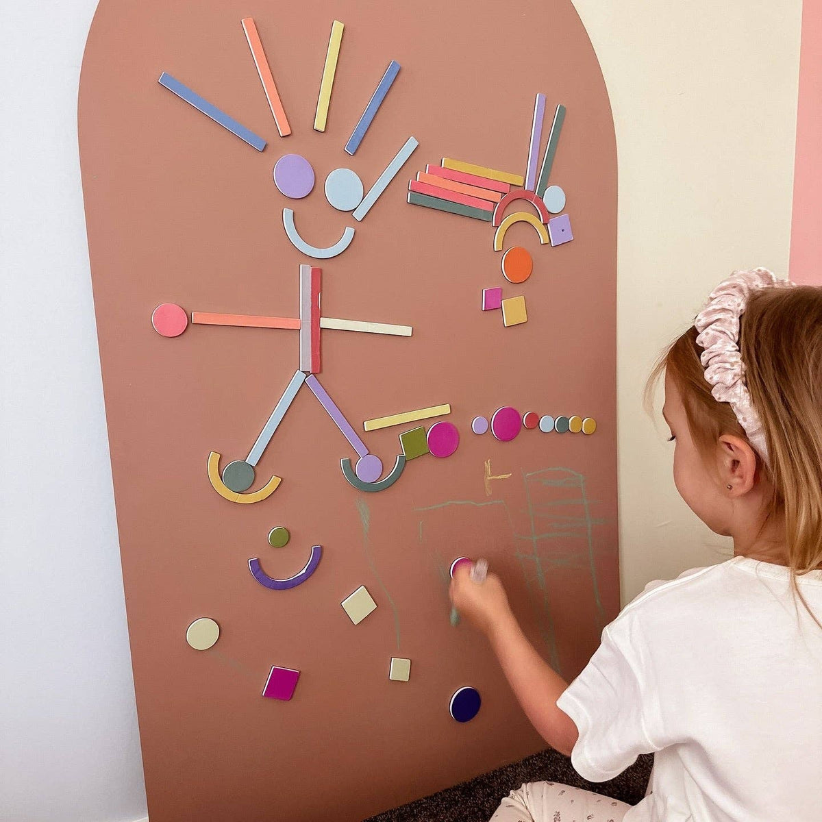 A young child wearing a white t-shirt and a pinkish headband is interacting with a large, arched, terracotta-colored magnetic board. The board is covered with colorful geometric shapes and stickers, and the child is reaching for a pink piece.