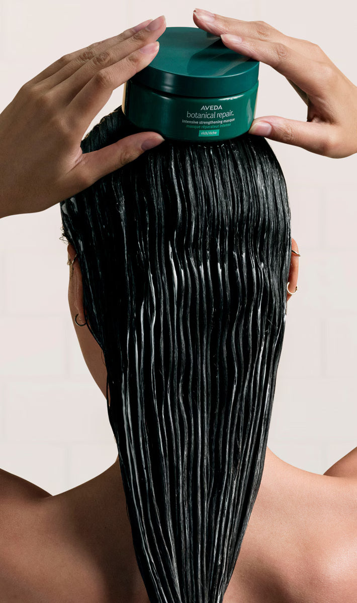 A person with long, dark, glossy hair applies or spreads a product from a dark green jar on their scalp, shown from behind against a light neutral background.
