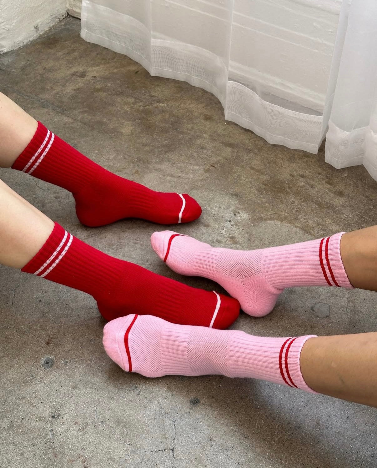 Red and pink socks with white stripes worn by two people on a concrete floor.