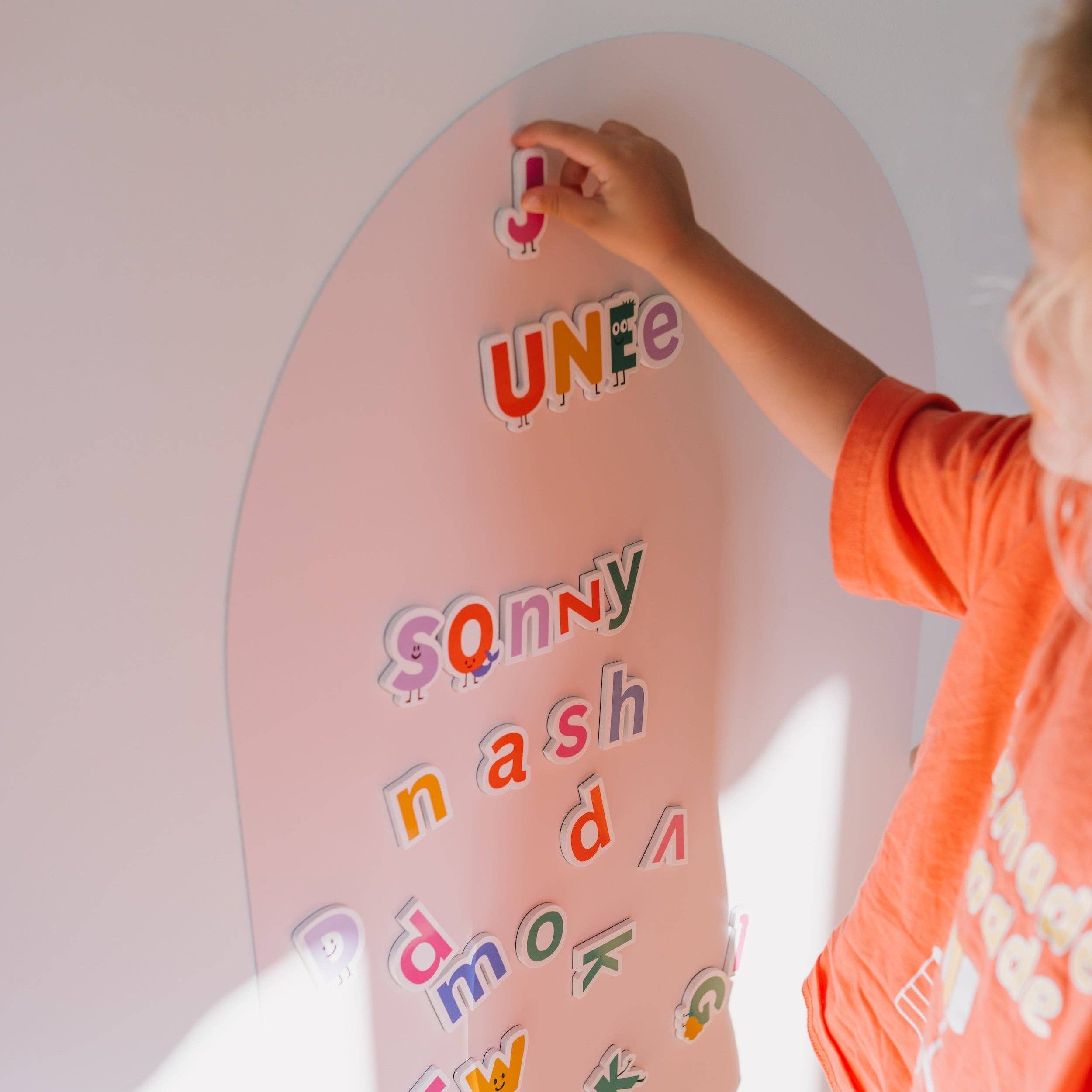 A child in an orange shirt is placing colorful magnetic letters on a pink arch-shaped board against a white wall.