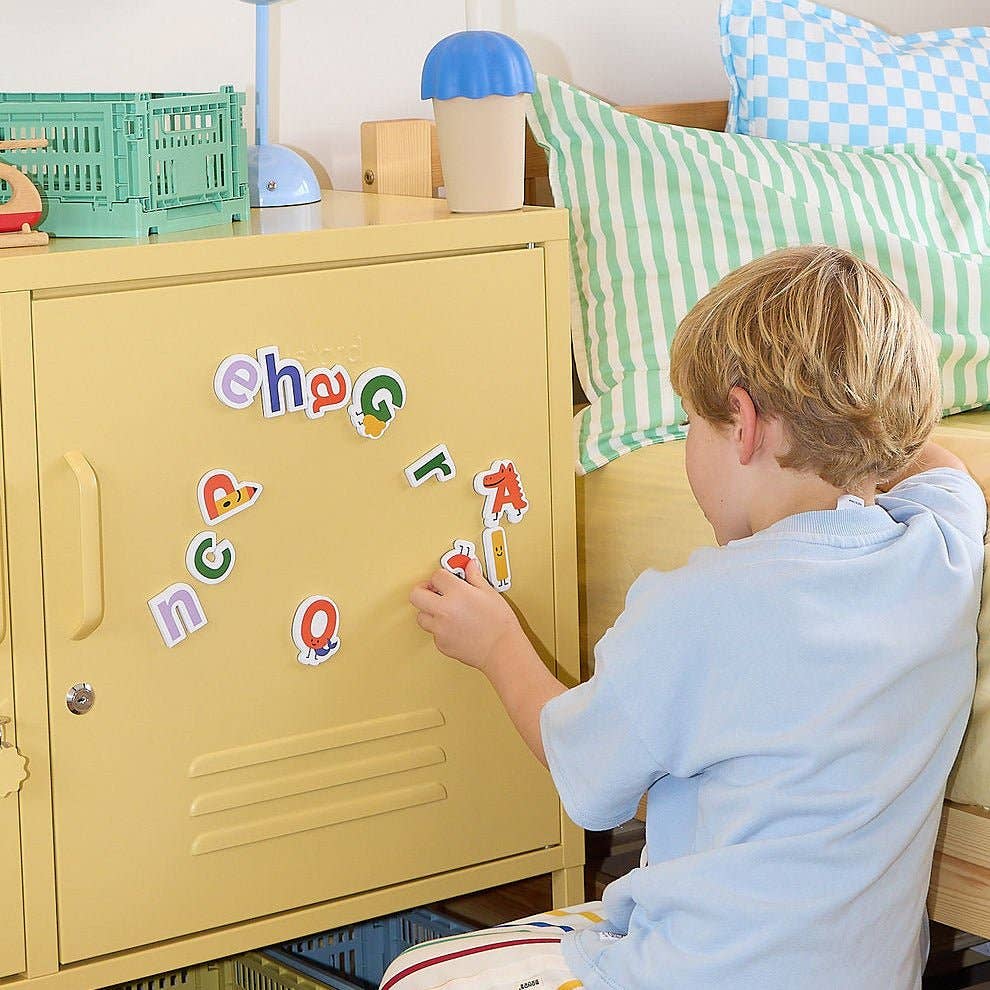 Child playing with alphabet magnets on a yellow locker in a room with green and blue decor.