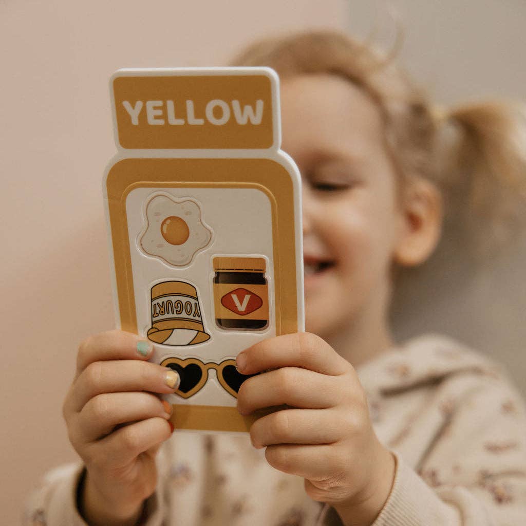 A young child holds up a yellow color identification card with stickers, with the child's face blurred in the background.