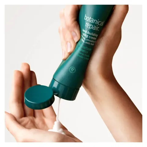 A person holds a dark teal bottle of botanical repair hair-building hair cream and squeezes white cream into their palm against a clean off-white background.