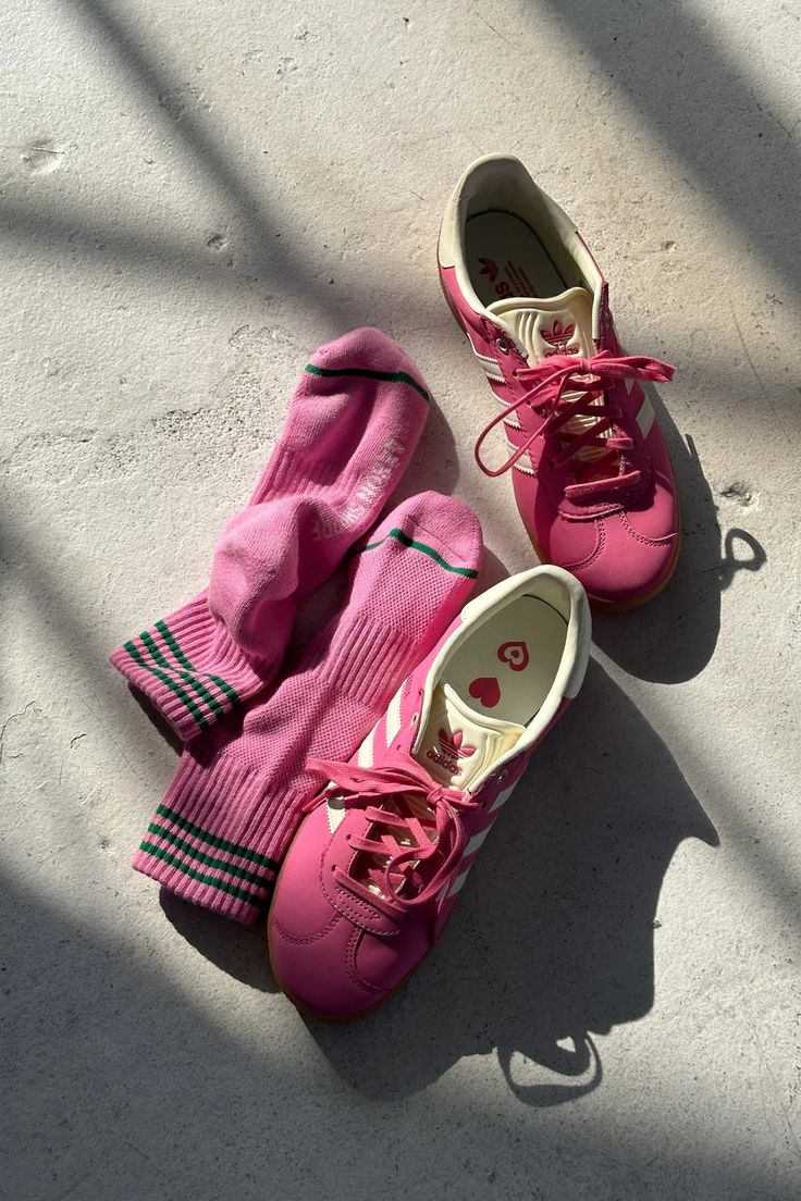 A pair of bright pink athletic sneakers and matching pink crew socks with black and green stripes, arranged on a sunlit concrete surface.