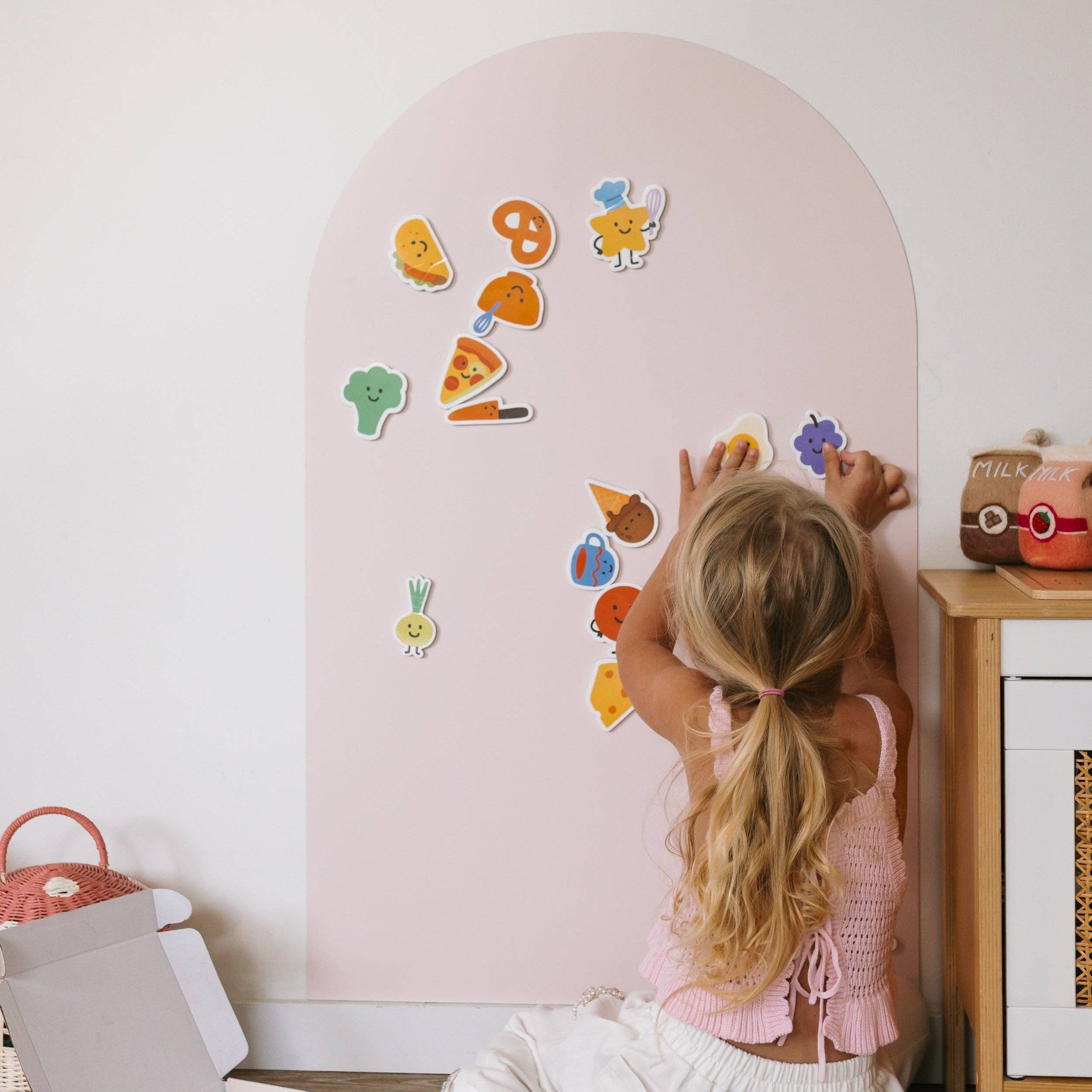 A young girl with blonde hair in a ponytail is seen from behind, interacting with colorful cartoon food and vegetable stickers on a light pink arch-shaped magnetic board. The scene is set in a bright, modern child's room with a wooden side table and toys nearby.