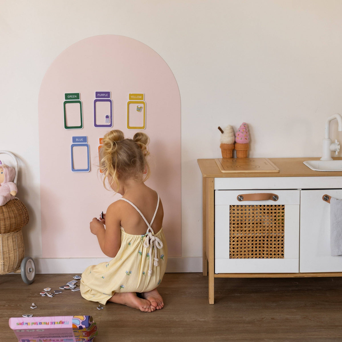A young child with blonde buns sits on the floor in a play kitchen setting, assembling small toy pieces. The room features a pastel pink arched wall with colorful toy organizer cards, a wooden and white play kitchen cabinet, and toy accessories.
