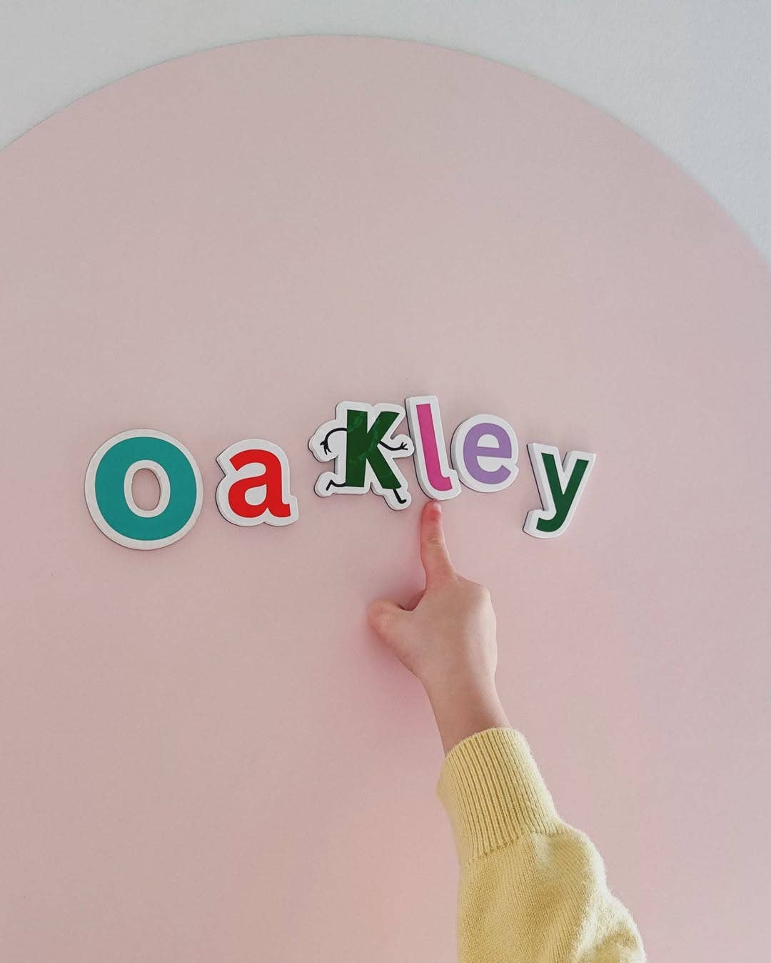 A child's hand wearing a yellow knit sleeve points to the letter 'K' in the word 'Oakley' spelled out with colorful magnetic letters on a pastel pink circular board.