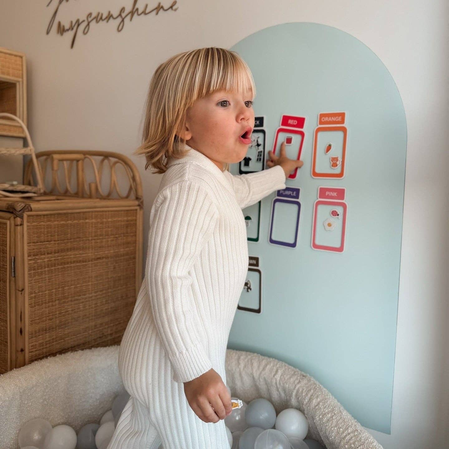 Child interacting with a colorful wall chart in a room with wicker furniture and balloons.