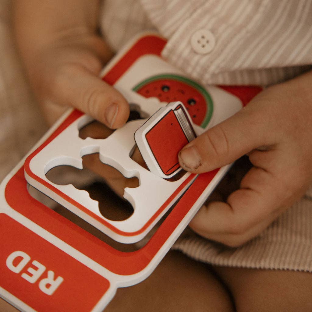 A close-up of a child's hands holding and playing with a red and white wooden shape-sorting toy board, featuring a watermelon slice piece and the word 'RED' on the board.