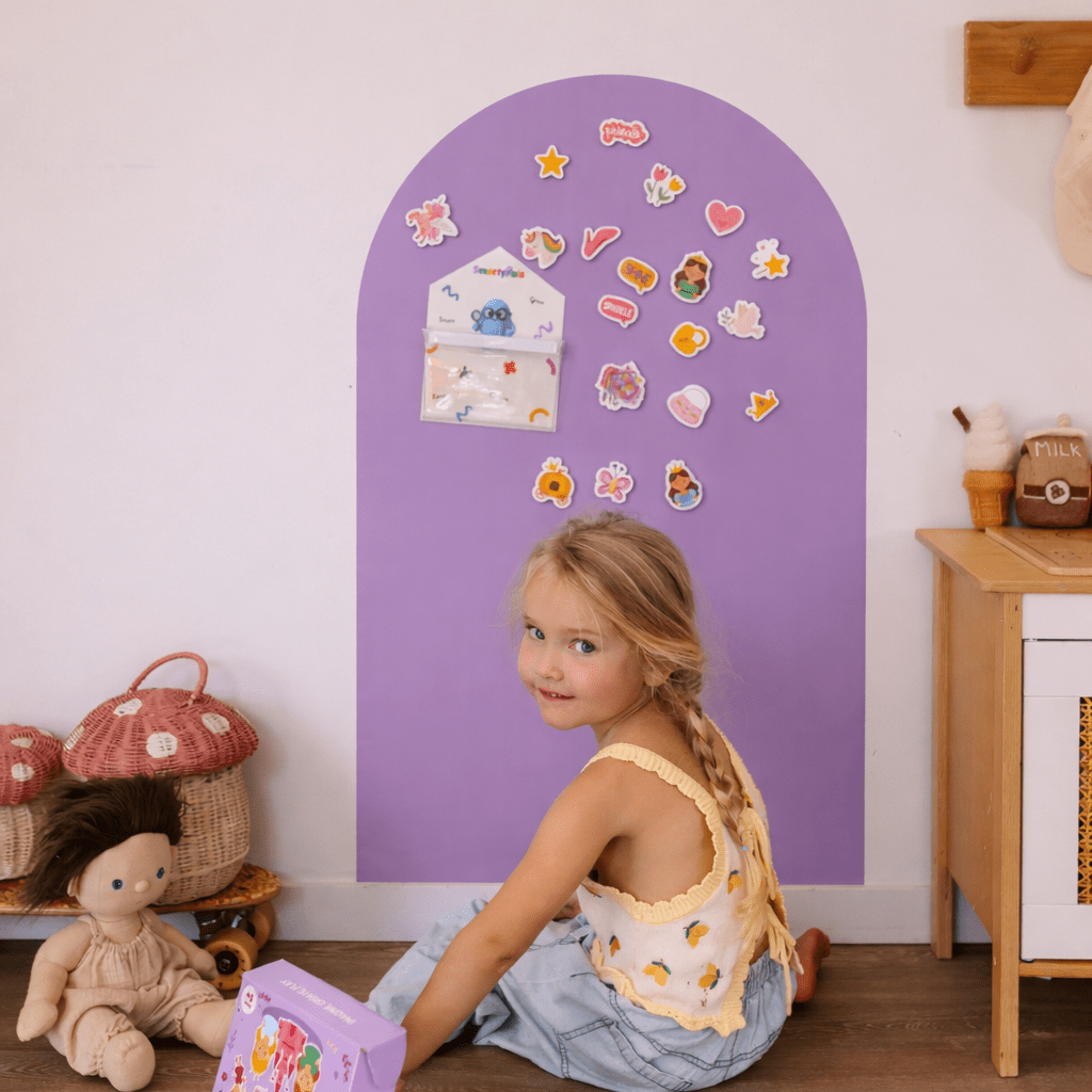 Child sitting on the floor in front of a purple arch with decorative stickers, with toys and a wooden cabinet in the background.
