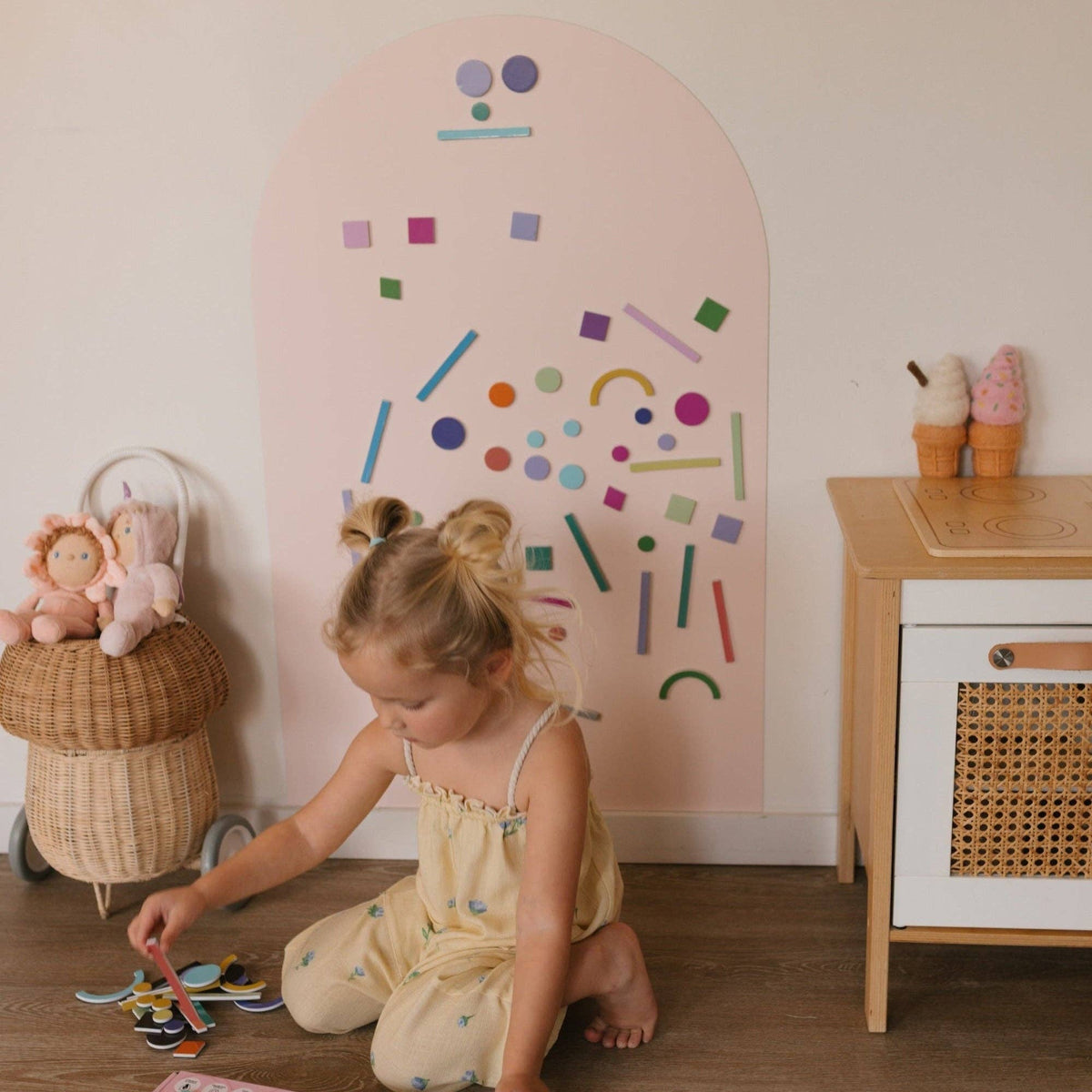 A young girl with twin buns sits on the wooden floor in a playroom, interacting with colorful geometric wall decals on a large arch-shaped pink board. She is wearing a pale yellow romper.