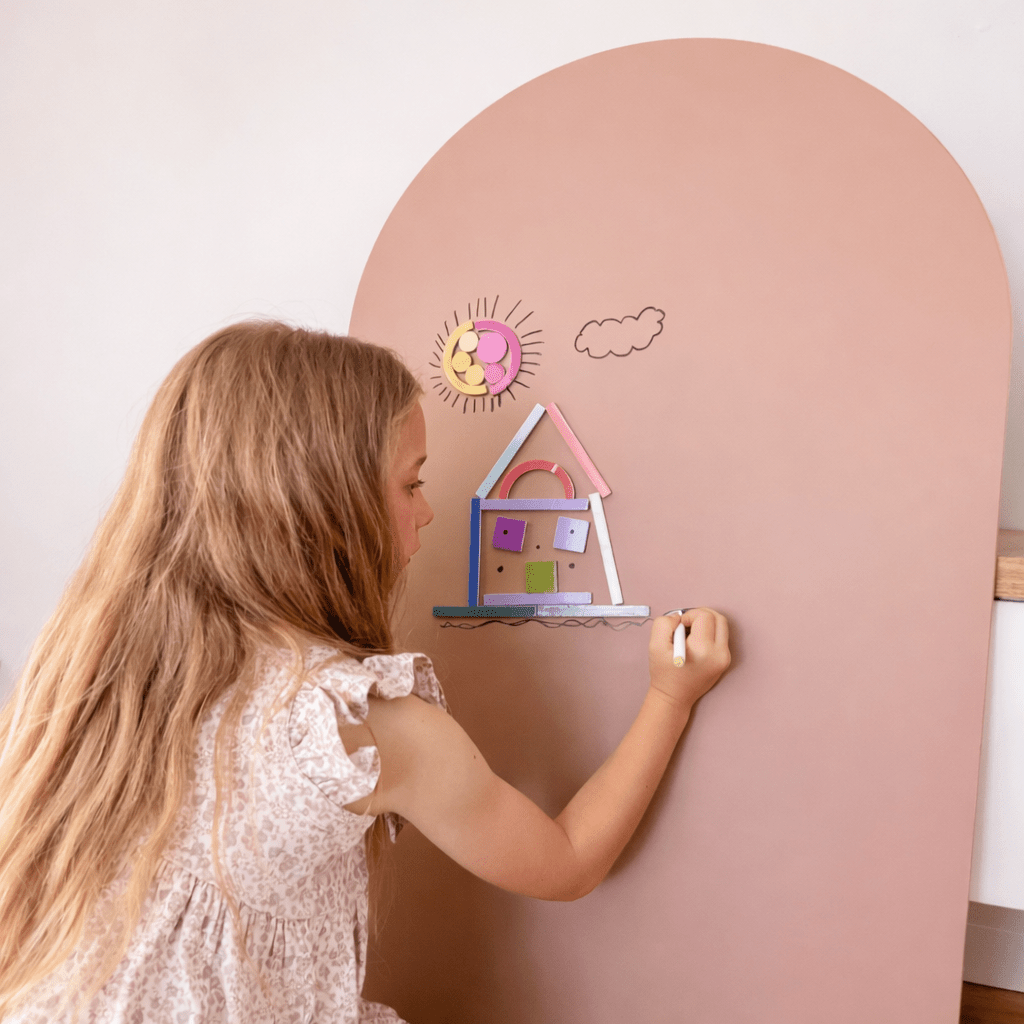 A young girl with long blonde hair is seen from the side, writing or drawing on a large dusty pink arch-shaped board. The board features a colorful hand-drawn house, sun, and cloud.