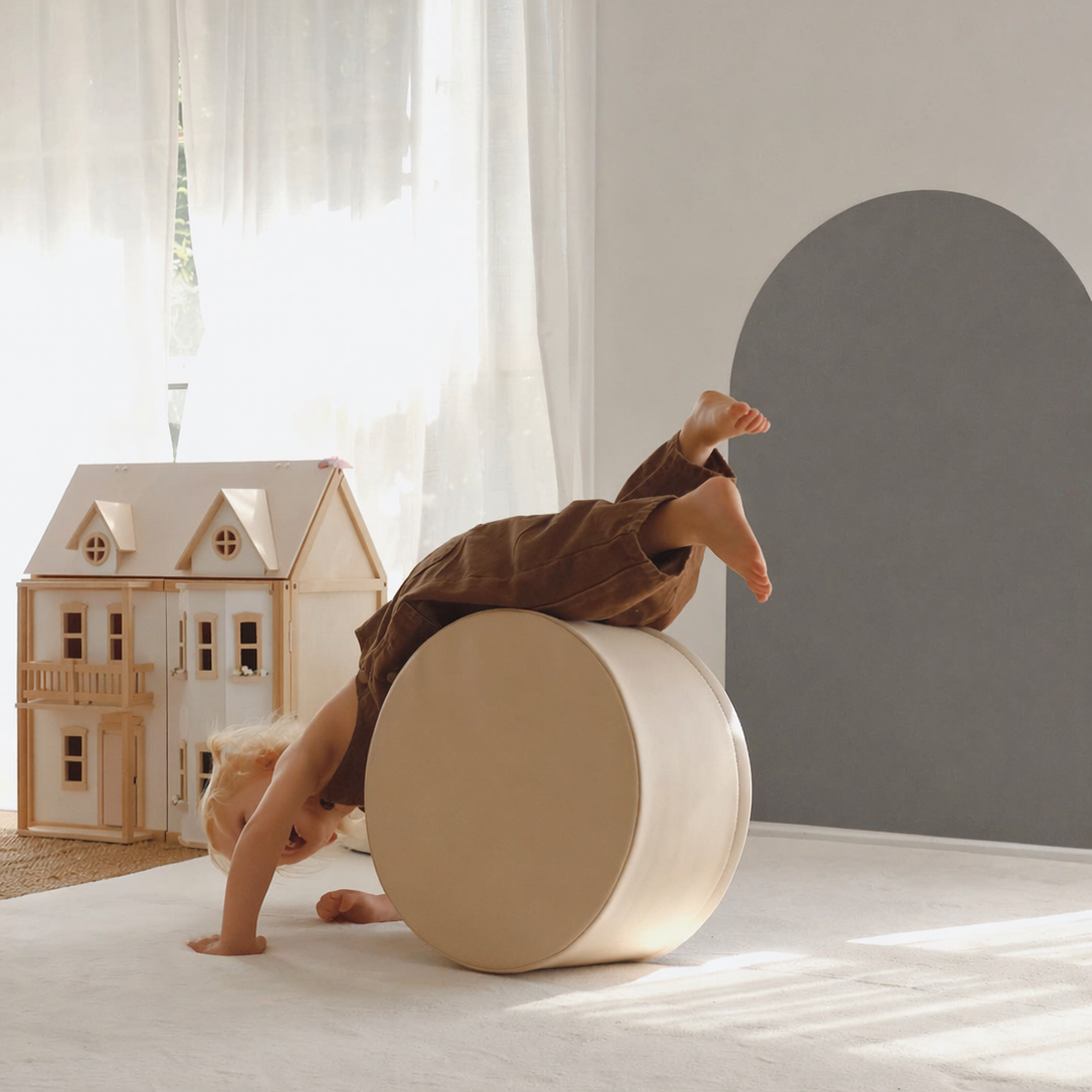 A child in brown clothing balances upside down on a large round beige exercise wheel in a bright, modern room with a dollhouse and a gray arched wall.