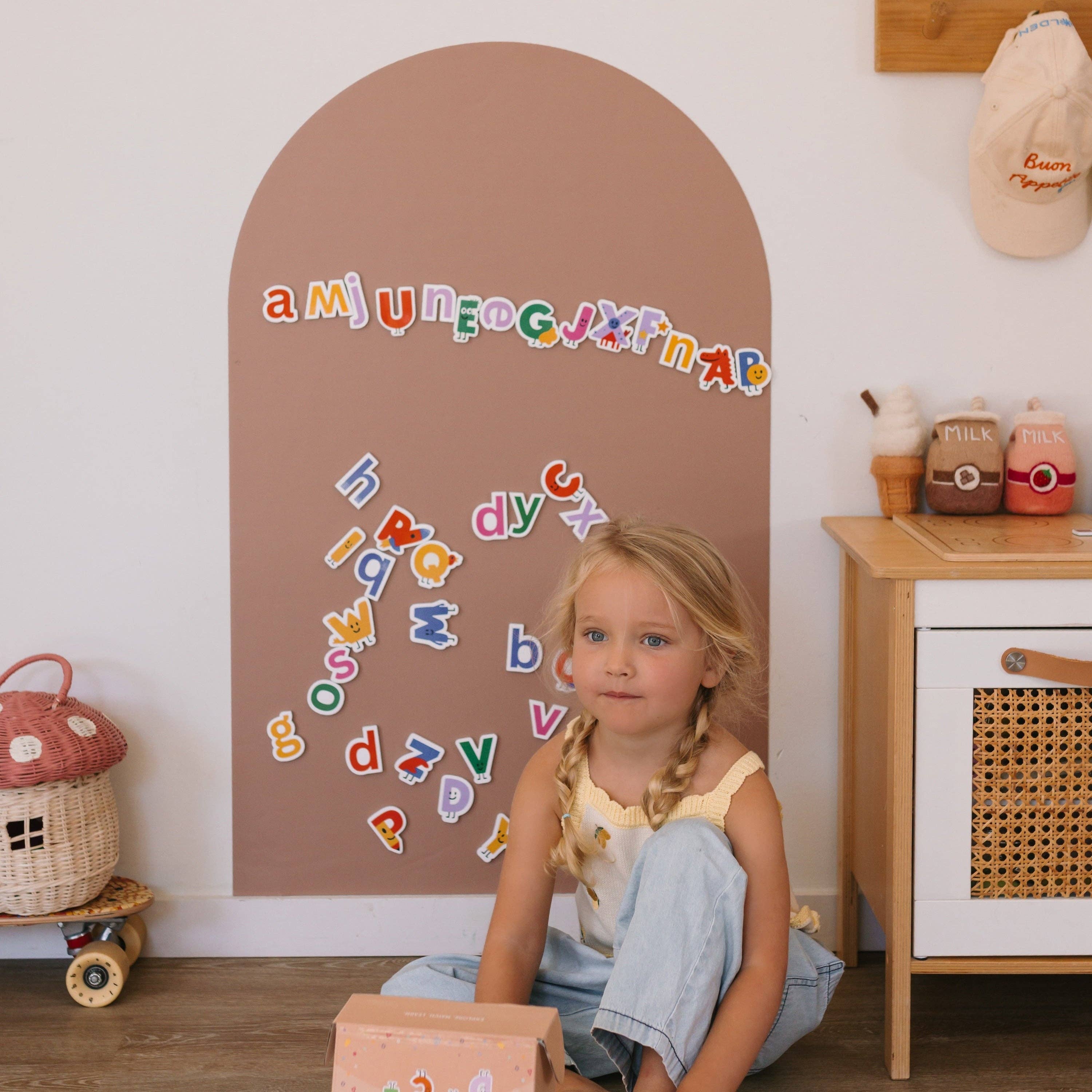 A young girl with blonde pigtails sits on the floor in a child's playroom, looking at the camera. In front of her is a large arched brown wall plaque decorated with colorful alphabet letters.