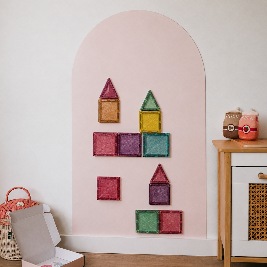 A pink arched wall display in a nursery or playroom setting, holding several colorful magnetic square and triangle building blocks.