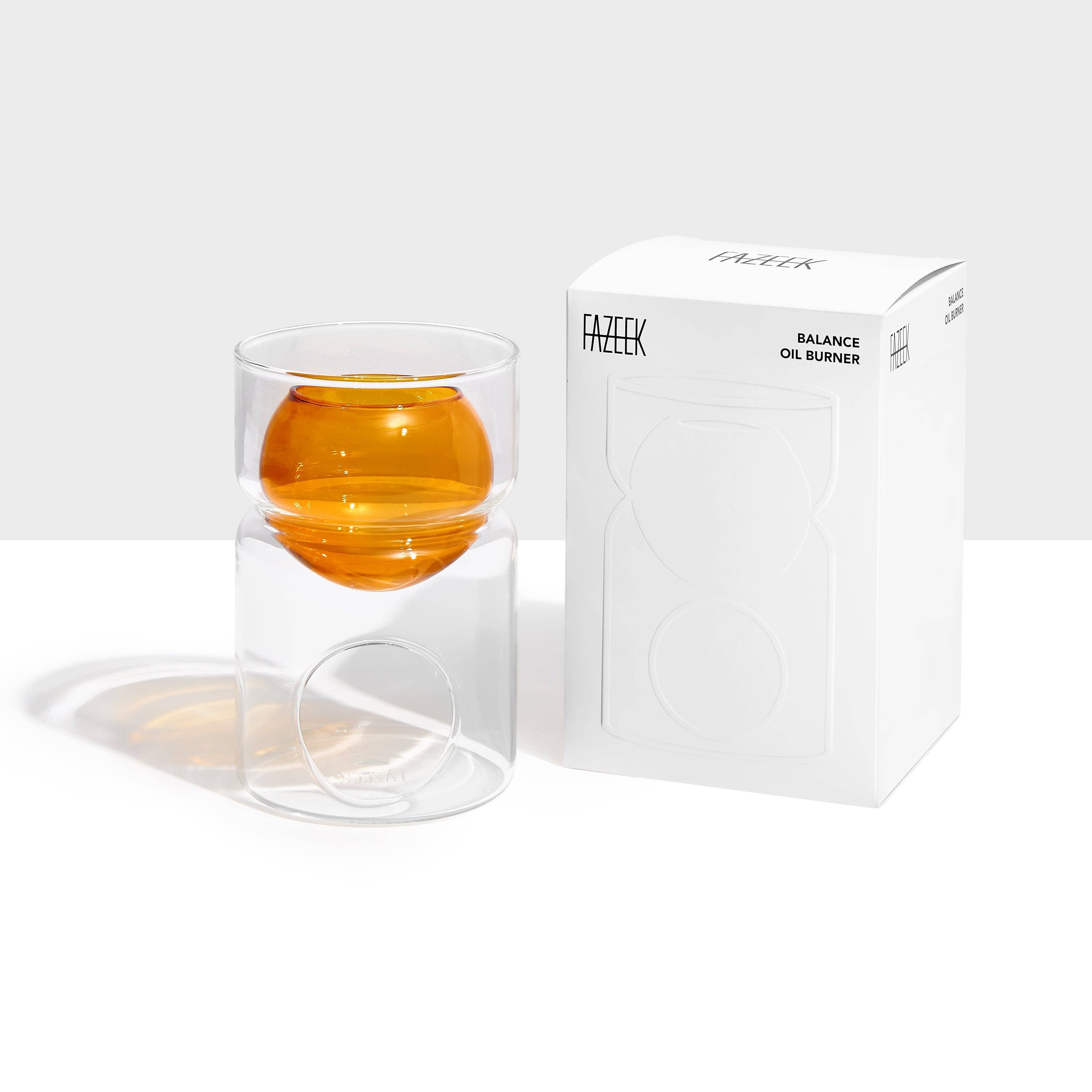 A clear glass balance oil burner with an amber-colored spherical oil chamber, shown next to its white branded packaging box on a clean white background.