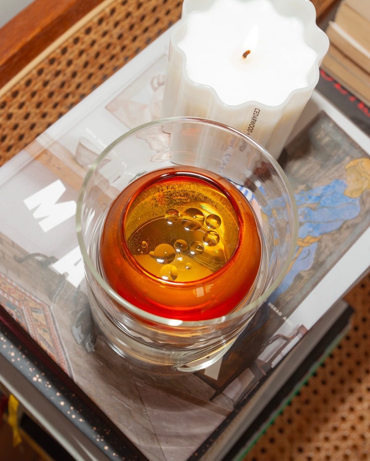 A clear glass candle holder filled with amber-orange wax, styled on top of books next to a white candle in a woven rattan tray.