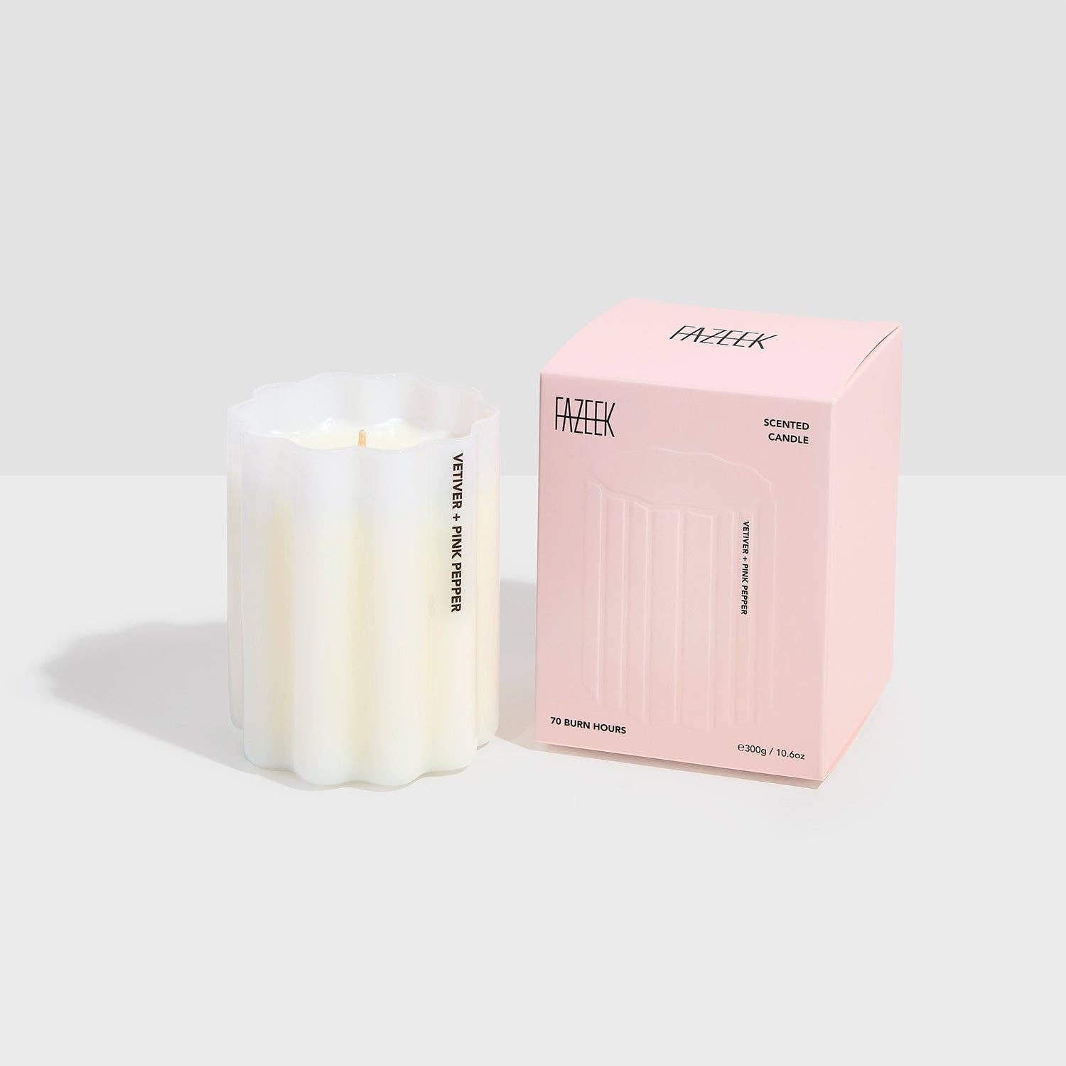 A white wax candle in a ribbed translucent glass vessel stands beside its light pink branded box on a clean light gray studio background. The box reads FAZEK and SCENTED CANDLE.