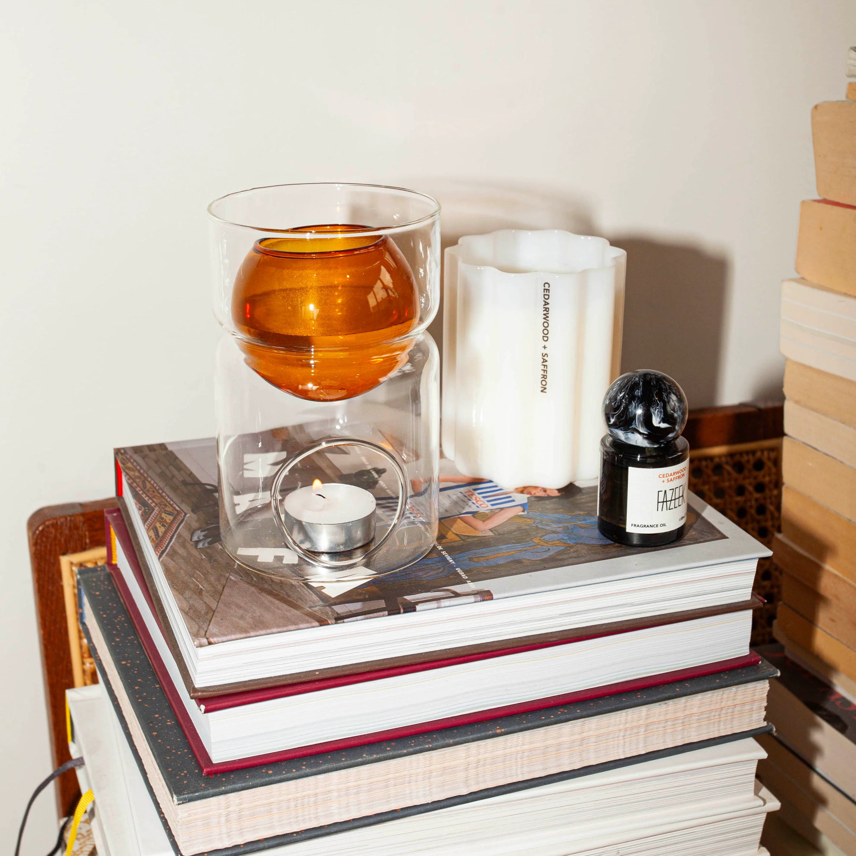 A modern glass double-walled candle holder with an amber-colored top bowl and a lit tealight candle inside, styled on top of a stack of books next to a white candle and a small black bottle.