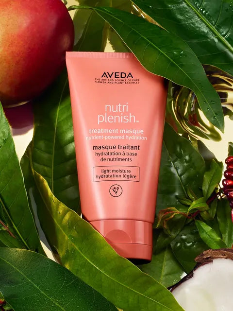 A tube of Aveda Nutri Plenish treatment masque in a coral pink color, nestled among green leaves, a red apple, and a slice of coconut.