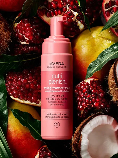 A bottle of Aveda Nutri Plenish styling treatment foam in a pink pump bottle, surrounded by fresh pomegranates, peaches, coconut, and green leaves.