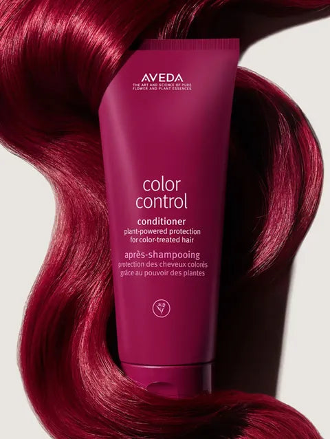 A tube of Aveda Color Control conditioner stands upright in front of flowing, glossy magenta hair on a light gray background.