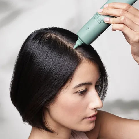 A woman with short black hair applies a light green hair product from a squeeze tube to the edge of her hair. The background is a soft, out-of-focus light gray.