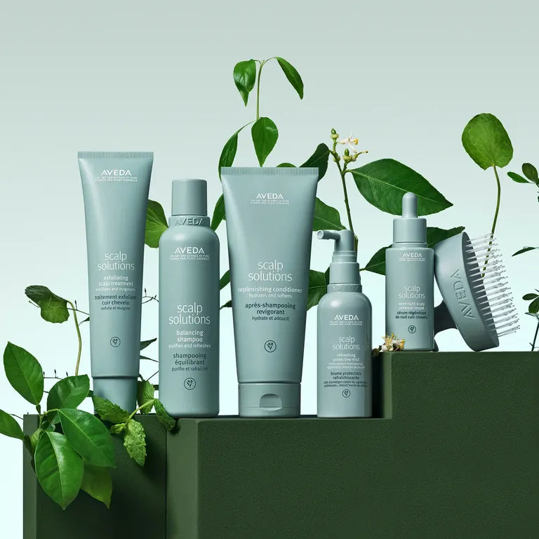 A set of Aveda Scalp Solutions hair care products in muted sage-gray packaging, arranged on a dark green pedestal with green leaves and small flowers around them. The products include a squeeze tube, a bottle, a larger squeeze tube, a pump bottle, a brush, and a comb.