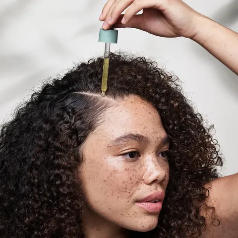 A close-up of a woman with curly dark hair applying a golden-yellow serum from a dropper onto her part line, set against a soft, light gray background.